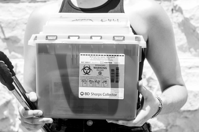 A person holds a pair of tongs and a box labeled "BD Sharps collector"