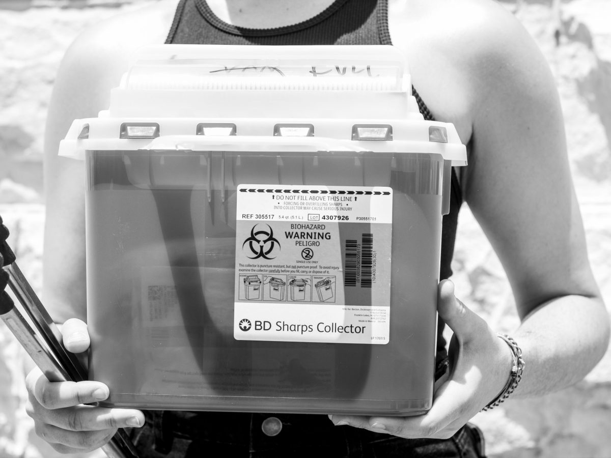 A person holds a pair of tongs and a box labeled "BD Sharps collector"