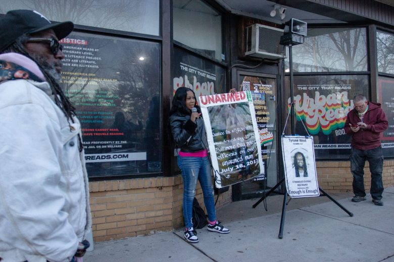 A photo of Tawanda West. She wears a black jacket and jeans. She us holding a large sign that says "unarmed" as she stands in front of Urban Reads bookstore.