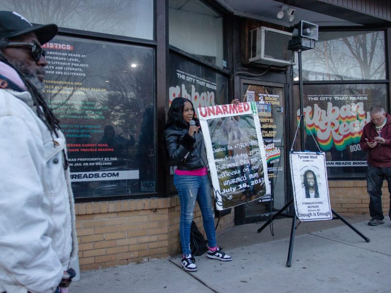 A photo of Tawanda West. She wears a black jacket and jeans. She us holding a large sign that says "unarmed" as she stands in front of Urban Reads bookstore.