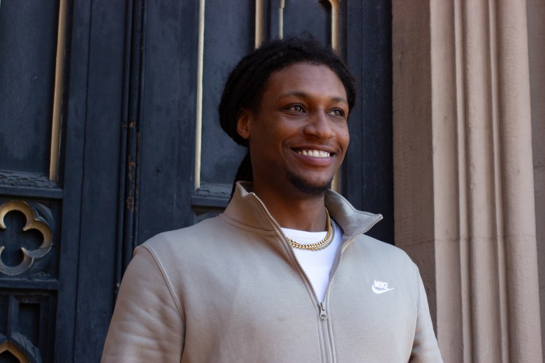 Greg Butler poses while wearing a tan jacket. He stands in front of a black door.