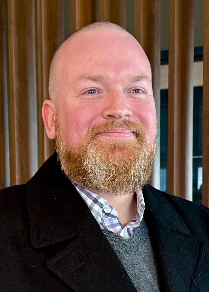 A man poses for a photo. He's weawring a blazer, a button down shirt, and a vest.