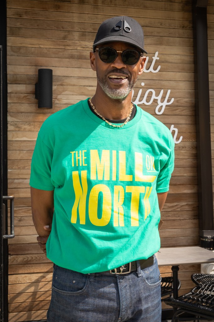 A man poses. He is wearing a black baseball cap and shades. He is wearing a green t-shirt that says "The Mill on North."