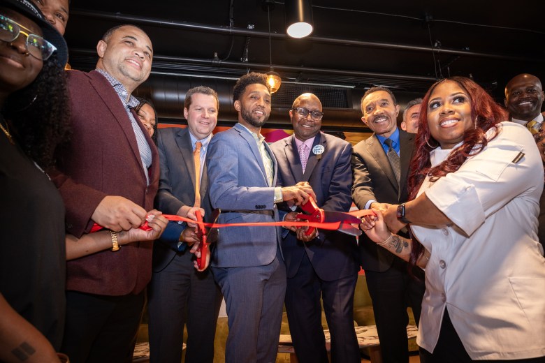 A group of people. Some are holding a large red ribbon. Two men are cutting the ribbon with large pairs of scissors.