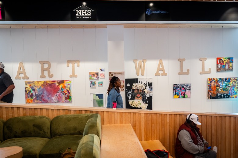 A woman walks past a white wall. The words "art wall" are spelled out on the wall with large letters. Brightly colored art is hanging on the wall.