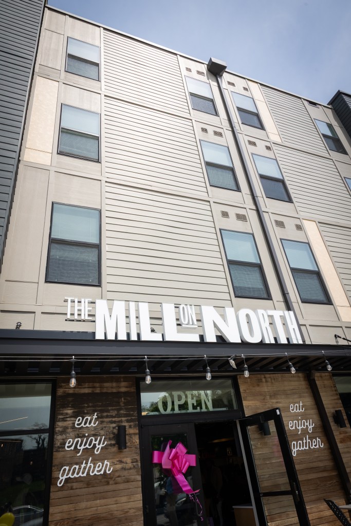 The exterior of a building. A sign reading "The Mill on North," is over the doorway. The words "eat enjoy gather" are on either side of the doorway.