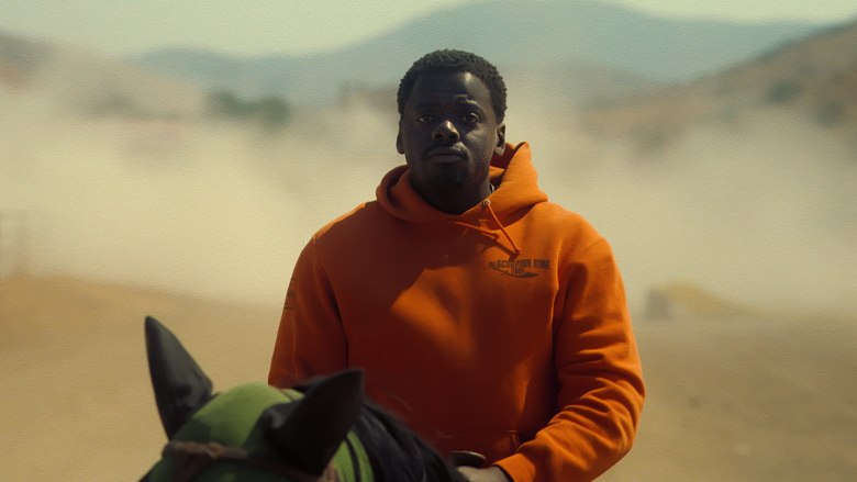 A still from the movie "Nope." Daniel Kaluuya as OJ rides a black horse. OJ wears a bright orange hoodie—promotional swag from the movie "The Scorpion King." OJ has a woried and resigned look on his face. The vast expansive mountains of California are behind OJ, out of focus.