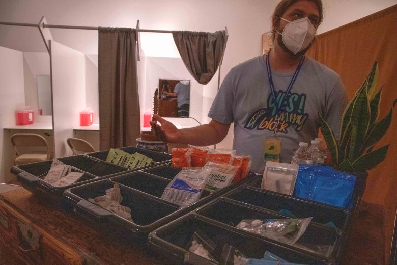 A harm reductionist stands in front of a safer use supplies. They are wearing a shirt that says "YES! ON MY BLOCK," in reference to advocacy to place overdose prevention sites in Baltimore. Behind the harm reductionist are the mock OPS room. The curtains are pulled revealing the lighting and mirrors in the OPS.