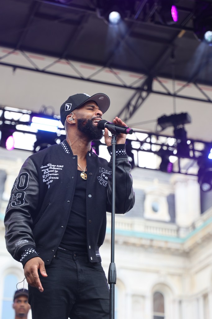 A man wearing a black hat, jacket, shirt and pants sings into a mic