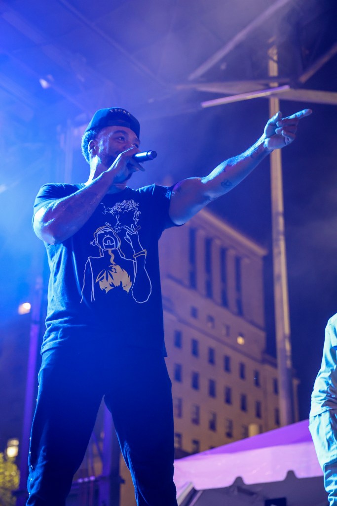 A man wearing a black hat and black shirt holds a mic and talks to a crowd.