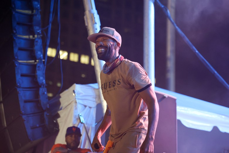 A man wearing a tan hat and t-shirt smiles while standing on stage.