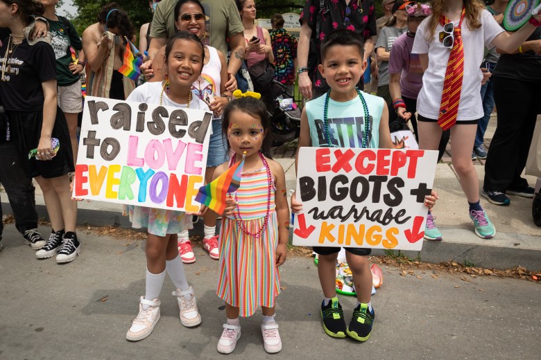 Siblings Malia, Laila, Luka watch the Baltimore Pride Parade with their mother while holding signs.