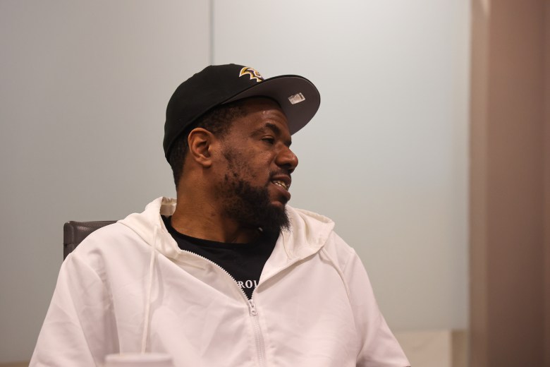 A Black man wearing a black baseball cap and a white shirt speaks.