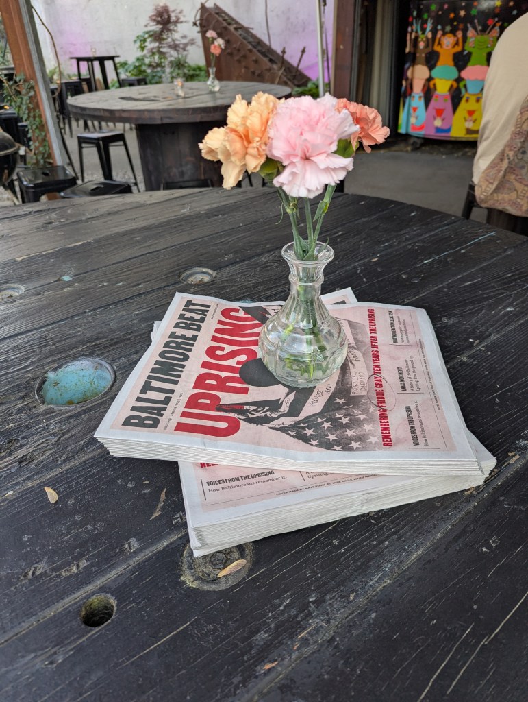 A pile of Baltimore Beat papers with a small vase of flowers on top of it.
