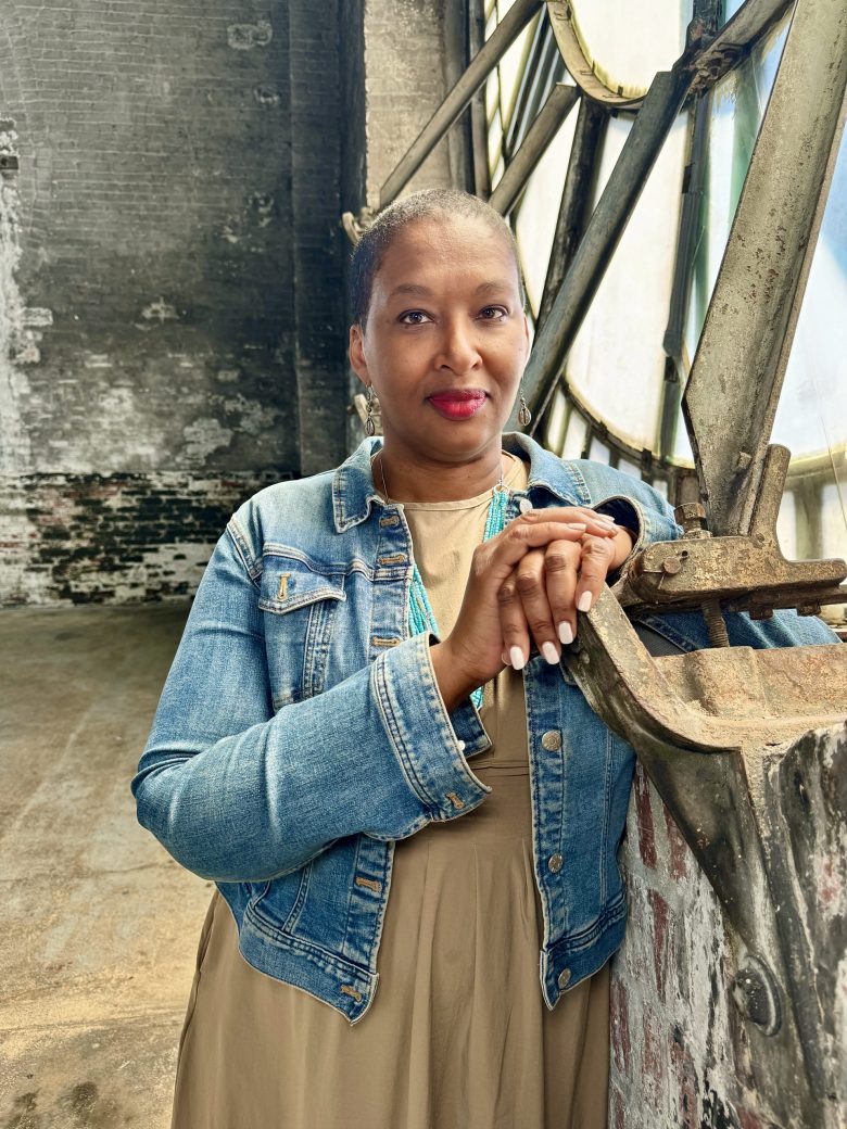 A Black Woman wearing a denim jacket and a brown top looks into the camera.
