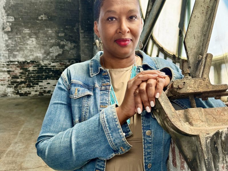 A Black Woman wearing a denim jacket and a brown top looks into the camera.