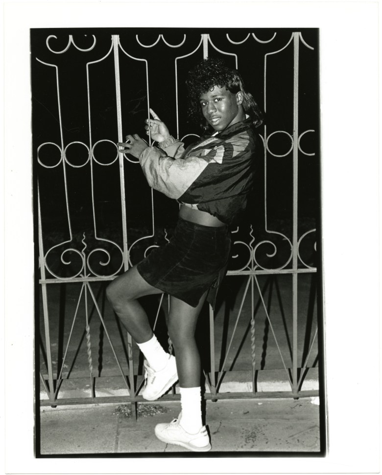 Portrait of a Trans-femme posed in front of an iron gate. The night scene features a full-length portrait of the subject, who playfully leans on the rungs of the gate. Her white socks glow from reflecting the flash, used by the photographer Joseph Kohl (1957-2002) to capture this nighttime scene.