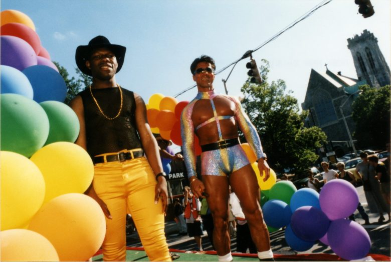 Two men in colorful pride fashions, including a sparkly kink-wear ensemble with hot pants. They appear to be standing on a Pride float adorned with rainbow balloons.