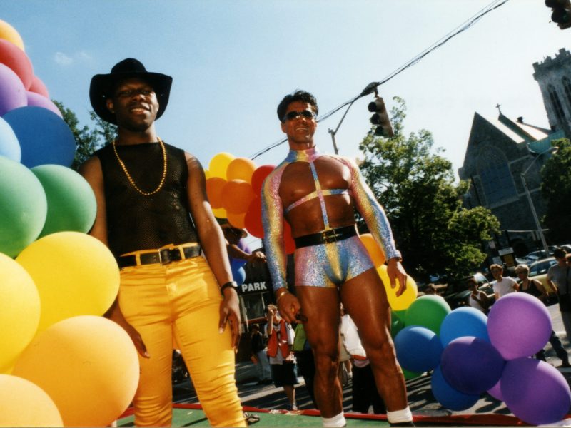 Two men in colorful pride fashions, including a sparkly kink-wear ensemble with hot pants. They appear to be standing on a Pride float adorned with rainbow balloons.