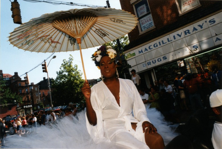 Scene from the Baltimore Pride Parade in 2000, on Charles street in the Mount Vernon neighborhood of Baltimore City. A femme in drag sits atop a fluffy white parade float in a silky white robe, white umbrella, and flowers in her hair.