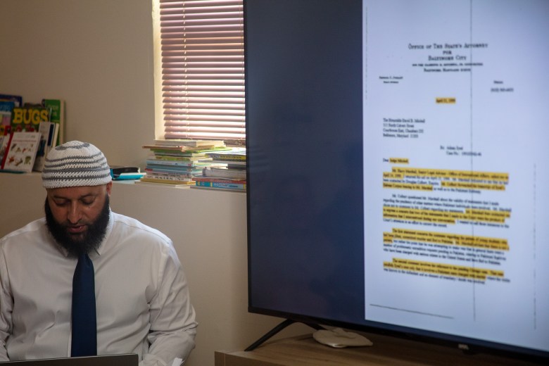 Syed looks down as he speaks. He sits next to a television screen with a court document on it.