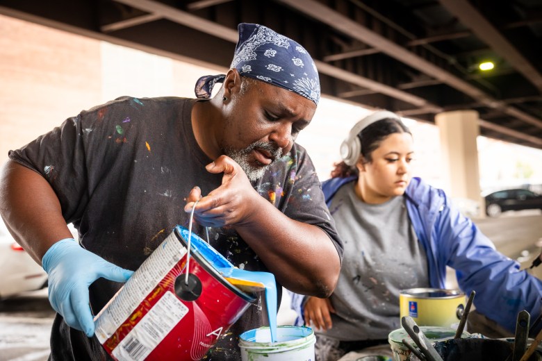 Shawn James pouring paint into a tub while his assistant Ariel Brown is behind him mixing paints.