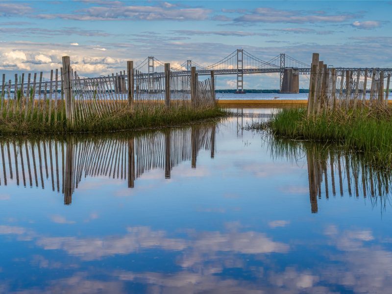The sky reflects off a body of water with a large bridge in the background.