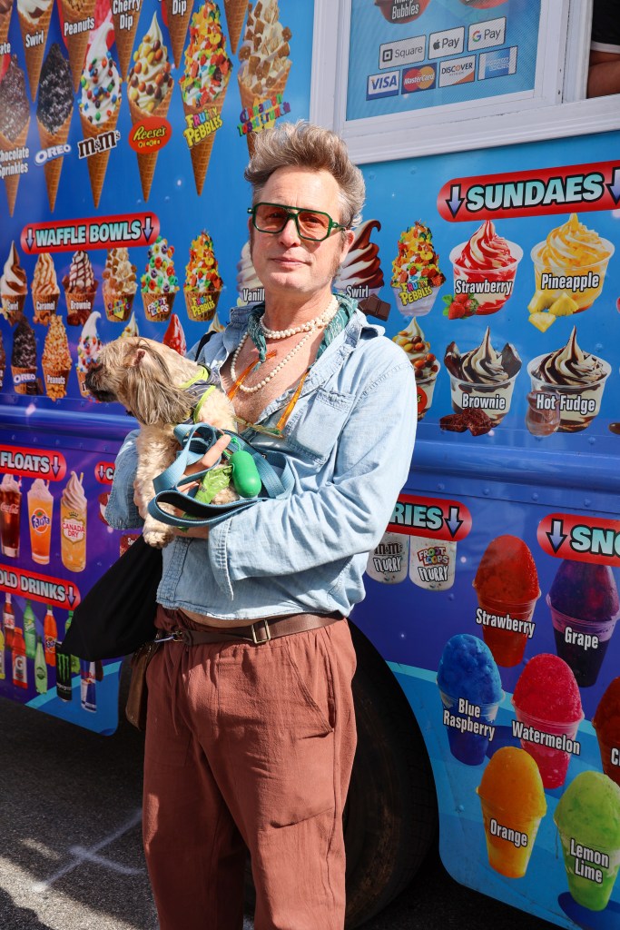 A man holding a small dog poses in front of an ice cream truck.