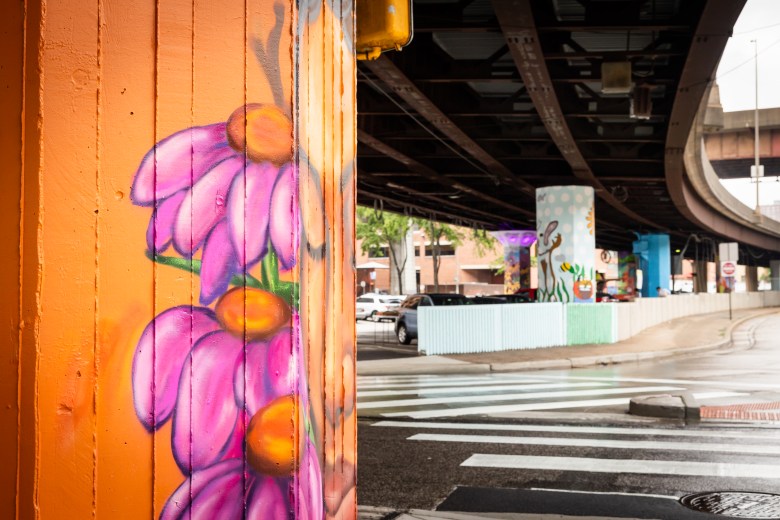 A flower painted on a pillar under the Jones Falls Expressway with several other murals visible in the background.