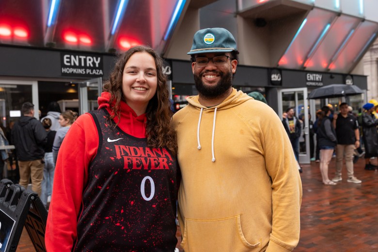 Two people pose for the camera. One person has on a green hat and a yellow hoodie. The other person has a red and black Indiana Fever jersey on over a red hoodie.