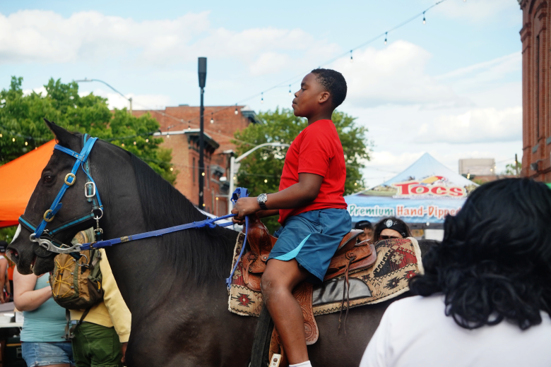 A child wearing a red shirt and blue shorts sits on a dark colored horse.