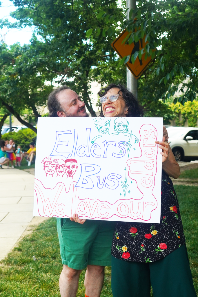 Two people smile happily while holding a large sign. The sign says: "Elders bus. We love our elders."