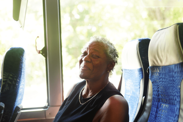 A person with brown skin and gray hair smiles from a bus seat.