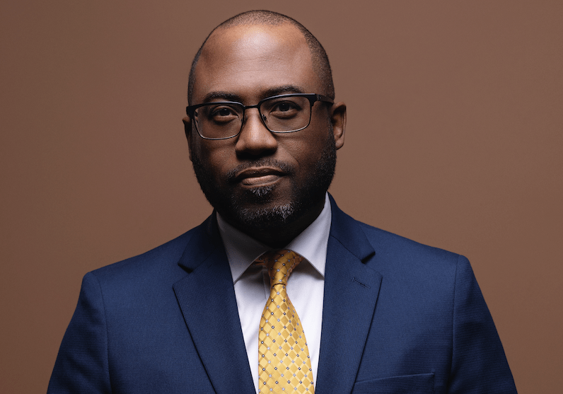 A photo of a Black man wearing a blue suit and a yellow tie.