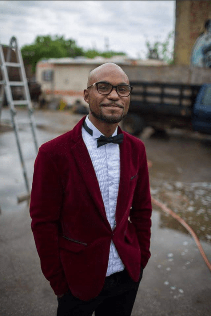 A man with brown skin poses for a photo. He is wearing a red velvet jacket, a white shirt, and a black bow-tie.